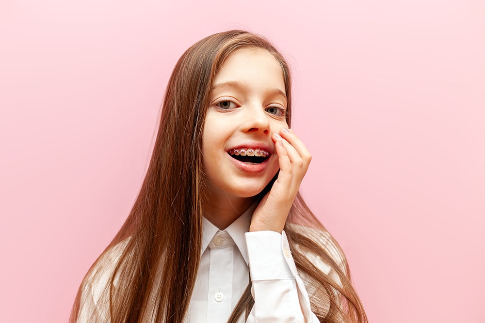 A girl with long hair showing her braces while touching her cheek, standing against a pink background