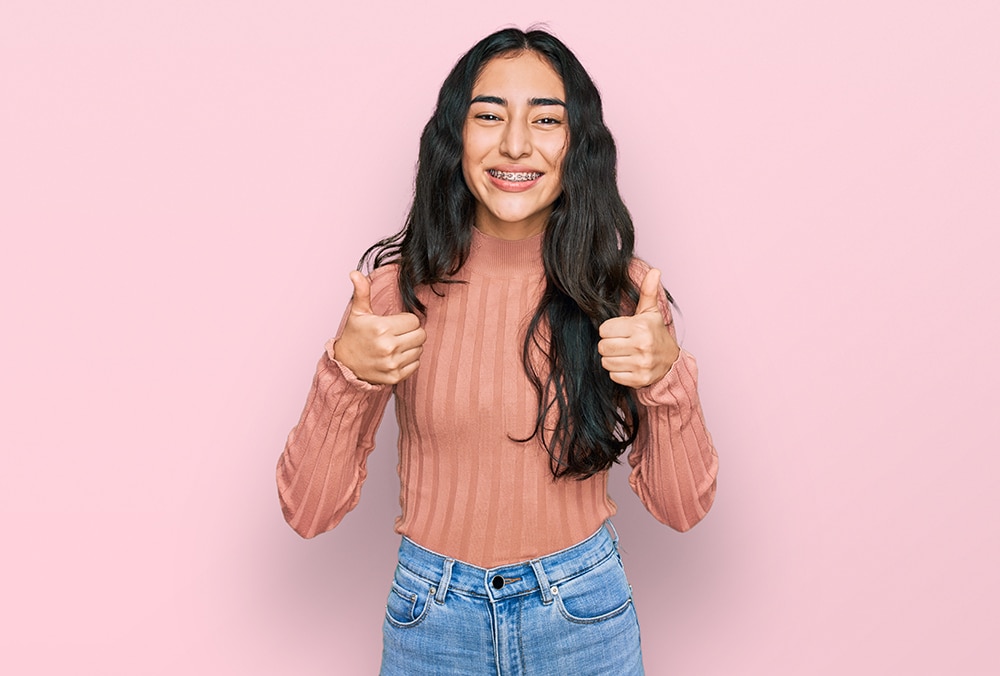 A teen girl with long dark hair, wearing braces, smiling and giving two thumbs up, standing against a pink background