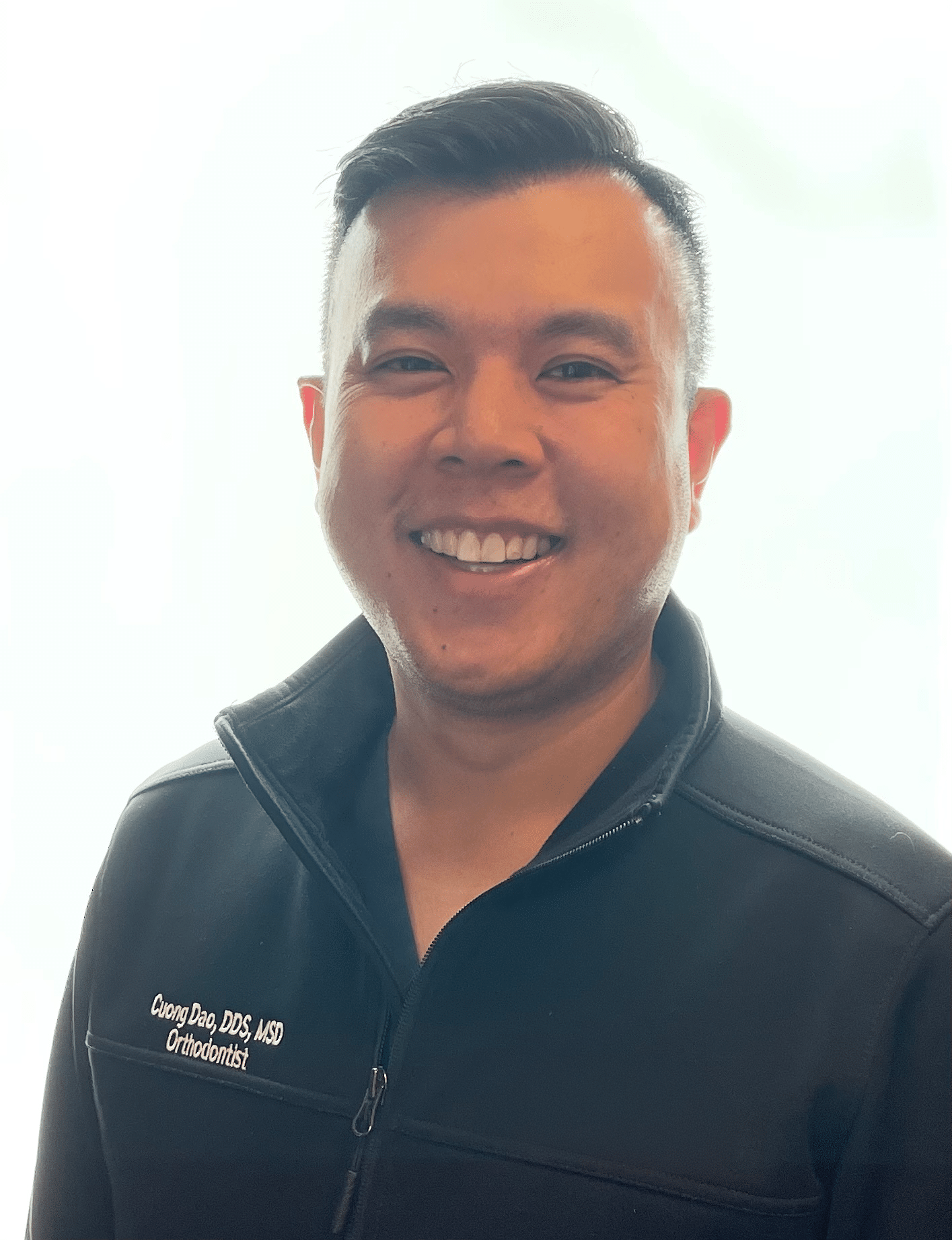 Headshot of Cuong Dao, an orthodontist, smiling in a black jacket embroidered with his name and title, against a light background